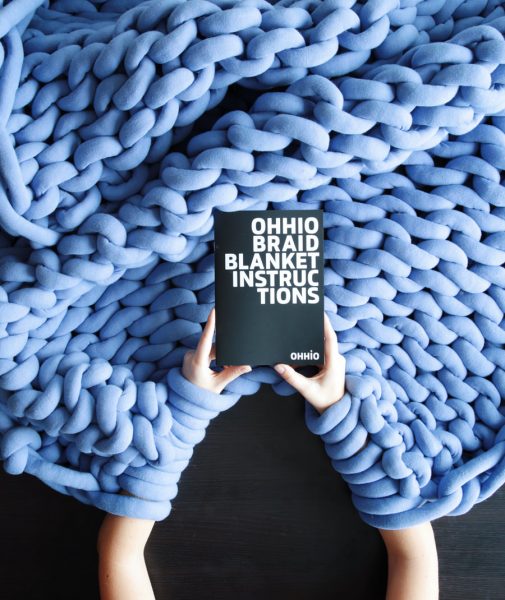 A shot from above of a woman's arms as she arm-knits a blanket. She's holding instructions for the project.