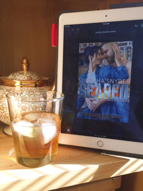 An amber colored drink next to a copy of Seared and a gilded teapot