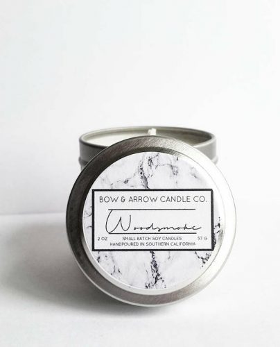 A soy candle in a metal tin. There's a marble print label on table with woodsmoke written in cursive.