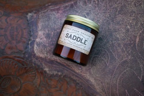 A candle in a deep reddish brown jar resting on a leather saddle.