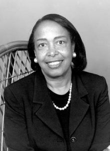Dr. Bath smiling at the camera, seated in a wicker chair wearing a dark suit and a white necklace and earrings.