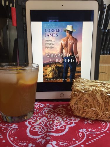 A copy of Wrapped and Strapped next to a glass filled with a muted orange liquid. There's a bandana underneath everything and a mini hay bale for decoration.