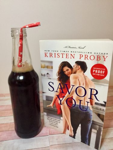 A drink in a glass coke bottle with a red straw. It's next to a copy of Savor You by Kristen Proby