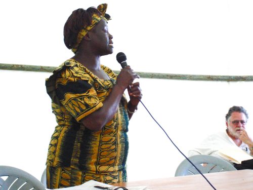 Wangari Maathai speaking into a microphone wearing a yellow and green print dress and head wrap