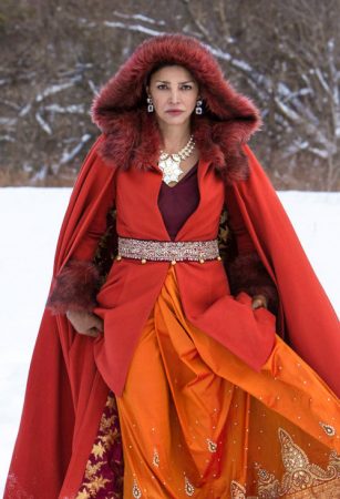 Chrisjen wears a long and flowing dress in reds and oranges. She has a jeweled belt and statement necklace. She is also wearing a red fur-lined cape with a hood.