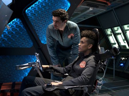 Naomi is in the pilot seat of a ship while Holden leans over her and watches a screen