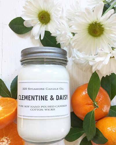 A white candle in a glass jar with a metal lid. The label is white with block, black letters. Around the candle is a bouquet of daisies and some clementines.