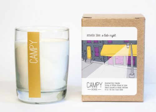 A white candle in an open glass jar. There's a mustard yellow stripe down the side that reads Campy. A small box sits next to the candle with the illustration of a street lamp outside of a bar.