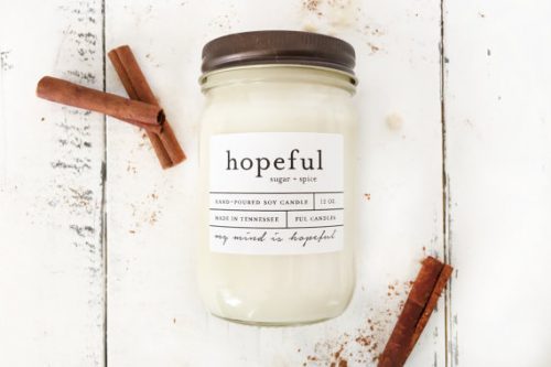 An off-white candle in a mason jar. It's resting on some white wooden boards with cinnamon sticks around it.