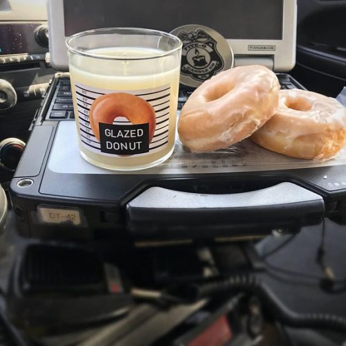 A clear candle jar with a label featuring a glazed donut. It sits on a police scanner with two donuts nearby.