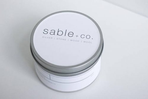 A small metal tin with a white candle. There's a white label on the outside and on top of the lid. The label is printed in thin, grey, minimalist lettering.
