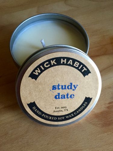 A white candle in a short metal tin. The name of the candle - Study Date - is printed in blue, type set letters on a parchment label.
