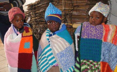 Three small children are wrapped up in hand knit blankets and hats