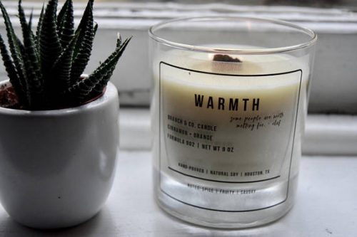 A clear jar with a white candle sitting in a sunny windowsill. The label text looks printed onto the glass and the candle has a square wood wick.