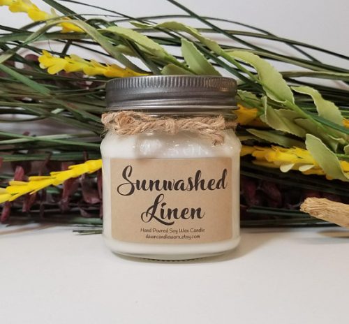 A white candle in a glass jar with a pewter lid and twine around the jar. The label is in parchment with black hand lettering. There's a bouquet of wild flowers in the background.