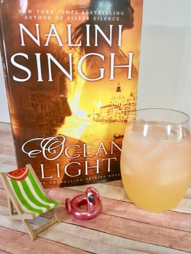 A cocktail in a short glass. The liquid starts off orange and lights to a yellowish pink. It's next to a copy of Ocean Light by Nalini Singh. A miniature beach chair and flamingo pool float are positioned nearby.