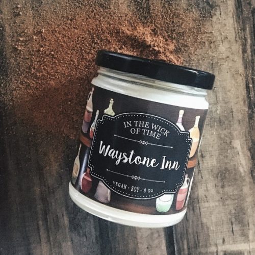 A white candle in a glass jar with a black lid. It's rest on a dark plank of wood. The label shows a wooden shelf with varies apothecary bottles.