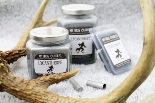 Mythos Candles in Lycanthrope. Two gray candles and a pack of gray wax melts on a furry, gray blanket. There's a deer antler and two silver casings positioned around them.