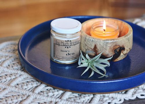 A white candle in a glass jar with a white lid. The candle is placed on a dark blue platter next to a wooden bowl and a small succulent plant.