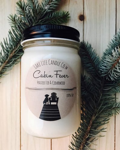 A large off white candle in a clear jar. It's resting on a wooden floor with pine needles around it.