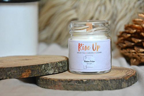 A white candle in a glass jar. It's resting on a wooden slab. The label is white and the Rise Up text is in a red to orange gradient.