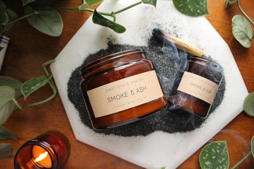 Two candles - one big and one small - in dark amber jars. They are resting on a pile of smoke and ash.