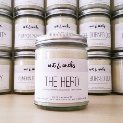 A white candle in a clear jar. The lid is a light metal and the candle is positioned in front of a wall of other candles.