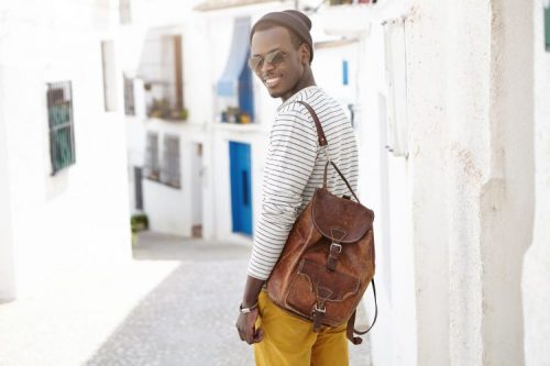 Outdoor shot of handsome happy young African American male traveler with leather backpack standing at concrete wall on narrow street while sightseeing in resort town during his summer holidays