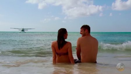 Becca and Garrett sit on the beach, the sea plane in the distance. The sea plane is very bored.