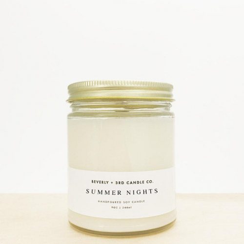 A white candle in a clear glass jar with a gold metal lid. The label is white and minimalist. It's resting on light wood in front of a white wall.