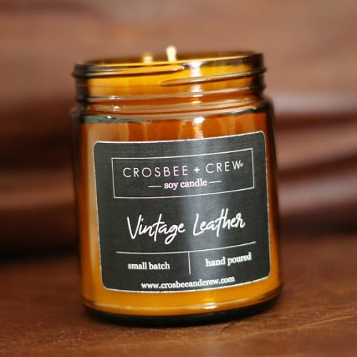 A lit white candle in an amber glass jar on a wooden table. The label is black with white text.