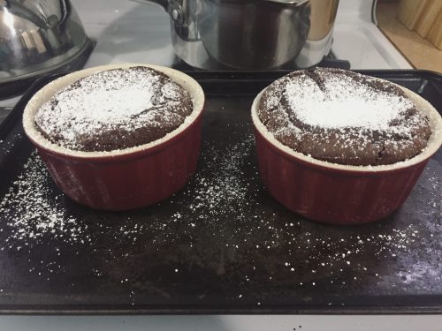 Two chocolate souffles in red ramekins. They're resting on a baking sheet and are dusting with sugar.