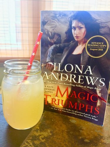 An ice cold yellow drink in a mason jar with a red straw next to a copy of Magic Triumphs