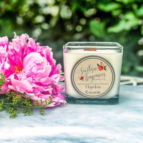 A white candle in a square glass jar. It's resting next to a bouquet of pink flowers.