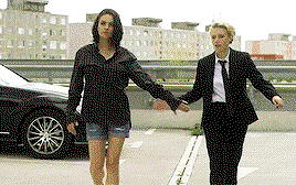 Kate and Mila, walking away from a car. Mila is in shorts and a dark plaid shirt, while Kate is in a tailored black suit with a tie. They're giving each other low five.