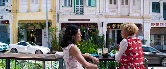 Rachel and Piek Lin talking at an outdoor cafe. Rachel is gesturing that she will not be intimidated by Nick's mother.