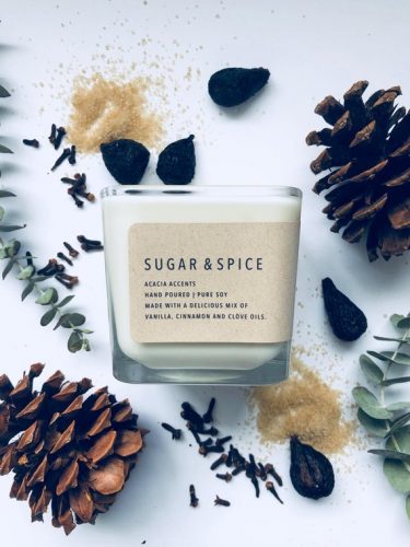 A white candle in a square jar. Around the candle are cloves, pinecones, and leaves.