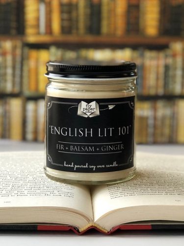 An off white candle with a black label. It's resting on an open book and displayed in front of a bookshelf.