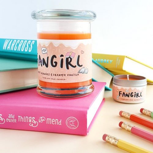 A bright orange candle on a light wood table. The candle is in a glass jar and is surrounded by colorful books and pencils.