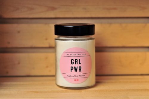 A white candle in a glass jar with a black lid. The circular label is a light pink.