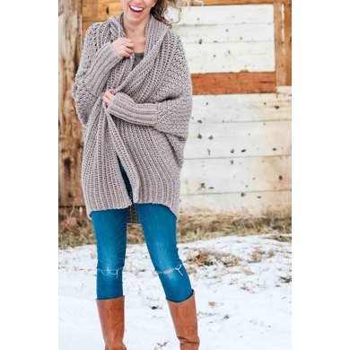 A woman models an oversized gray cardigan against a rustic background.