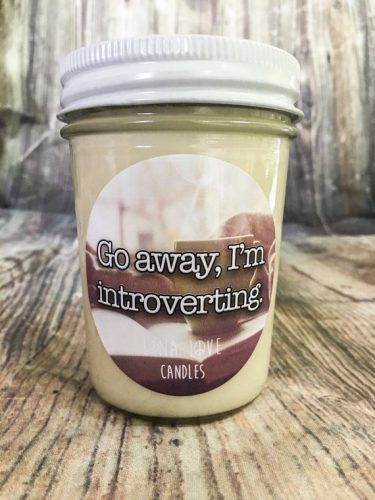 An off white candle in a clear jar in front of a wooden background. The label has a photo of books and a teacup on it.