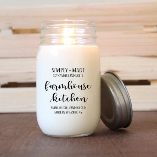 A white lit candle in a large glass jar. The candle is on a dark wood table in front of some light wooden slats.