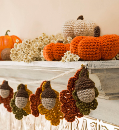 A rustic looking shelf is topped with crochet pumpkins. A garland made of crocheted acorns and leaves dangles from the bottom.