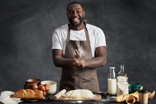 Cheerful African American male cook presents culinary talents participates in television cooking show wears apron stands at kitchen table makes dough for preparing buns isolated on chalk wall