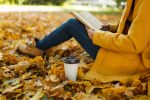 The woman in yellow coat jeans and boots sitting under the maple tree with a red book and cup of coffee or tea in fall city park on a warm day. Autumn golden leaves. Reading concept. Close up.