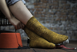 The feet of a model display socks that have a textured pattern that looks like the Daleks from Doctor Who. The socks are beaded to accent the pattern and are knit in a gold/bronze colored yarn. The model is perched on a stool