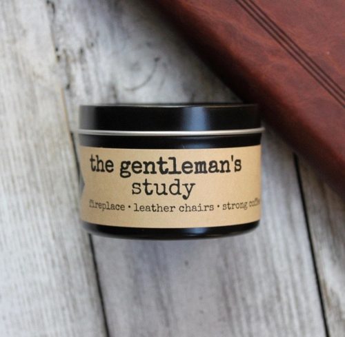 A candle in a black tin with a lid. It's on a worn wooden table next to a bound leather journal.