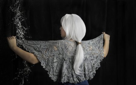 We see the back of a model wearing the Orichalcum shawl. It's a gray half-moon shawl with lots of lacework. The model has a bright white wig on, pulled back into a ponytail and is holding the edges of the shawl up so we can see the pattern clearly.
