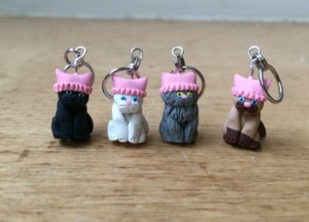 Four polymer stitch markers of cats wearing bright pink pussy hats. There is a Siamese cat, a longhair grey cat, a white cat and a black cat.
